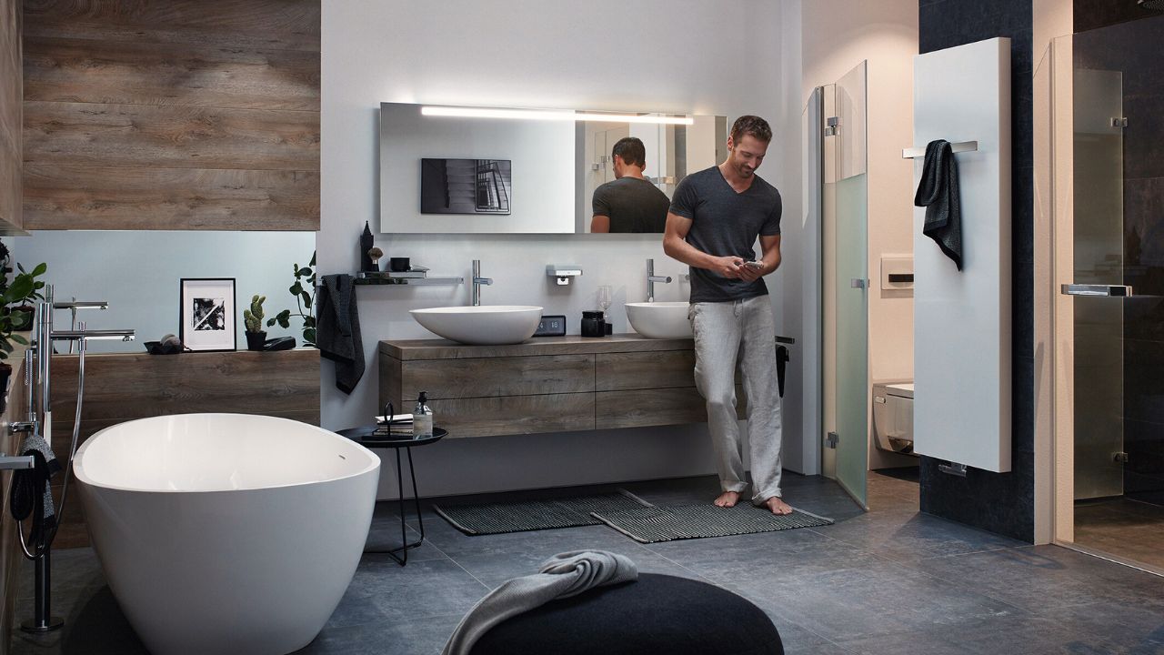 Mann ist barfuß und lehnt an Waschbecken im Badezimmer; er regelt über sein Handy die Termparatur der Fußbodenheizung