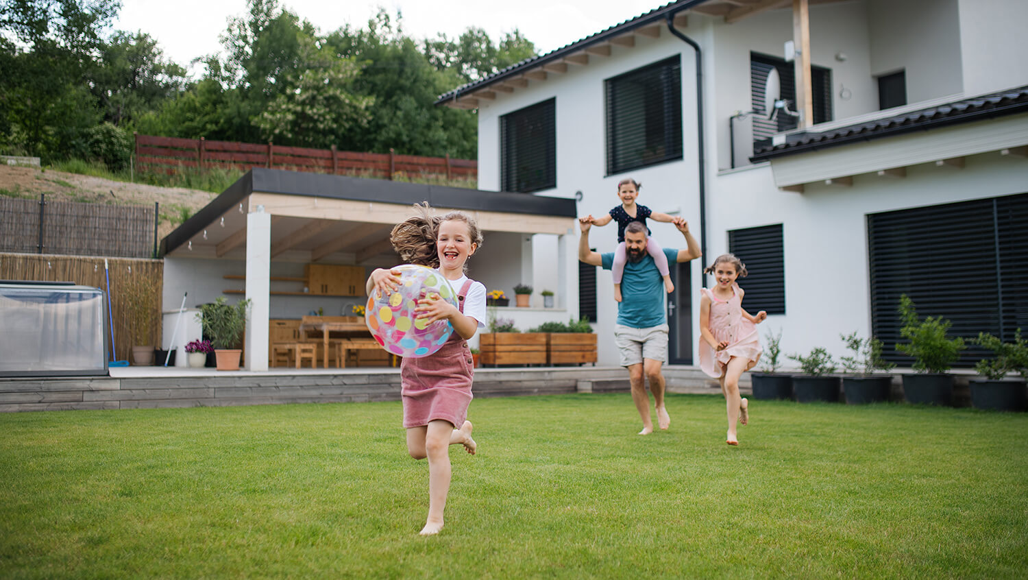 Ein Vater rennt mit seinen Töchtern auf einer  Wiese vor dem  Haus.