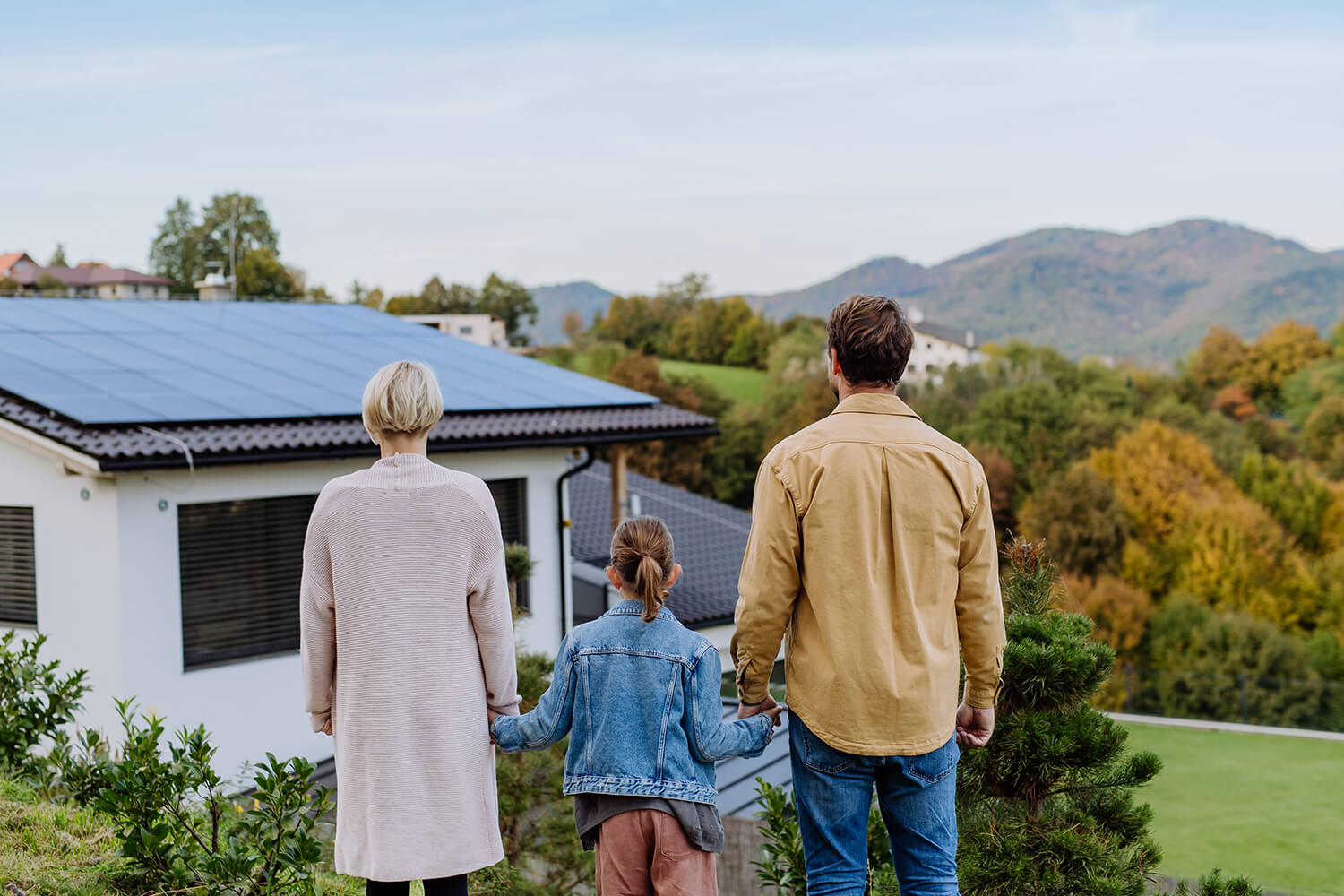 Eime Familie steht vor einem Haus mit einer Solaranlage auf dem Dach. Die Familie ist von hinten zu sehen. 