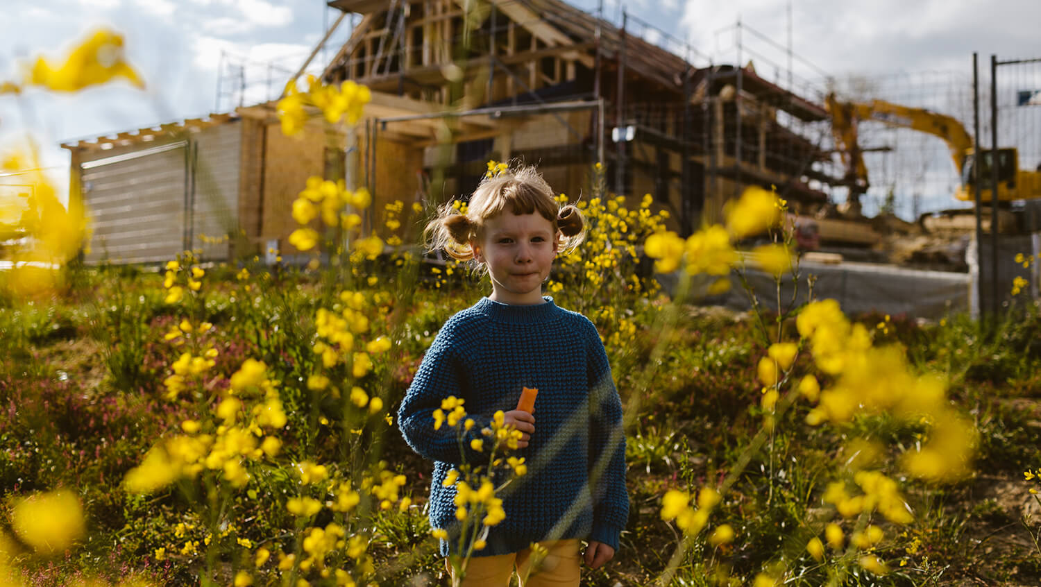 Ein Mädchen steht in einer blühenden Blumenwiese. Im Hintergrund ist ein Haus im Rohbau zu sehen.