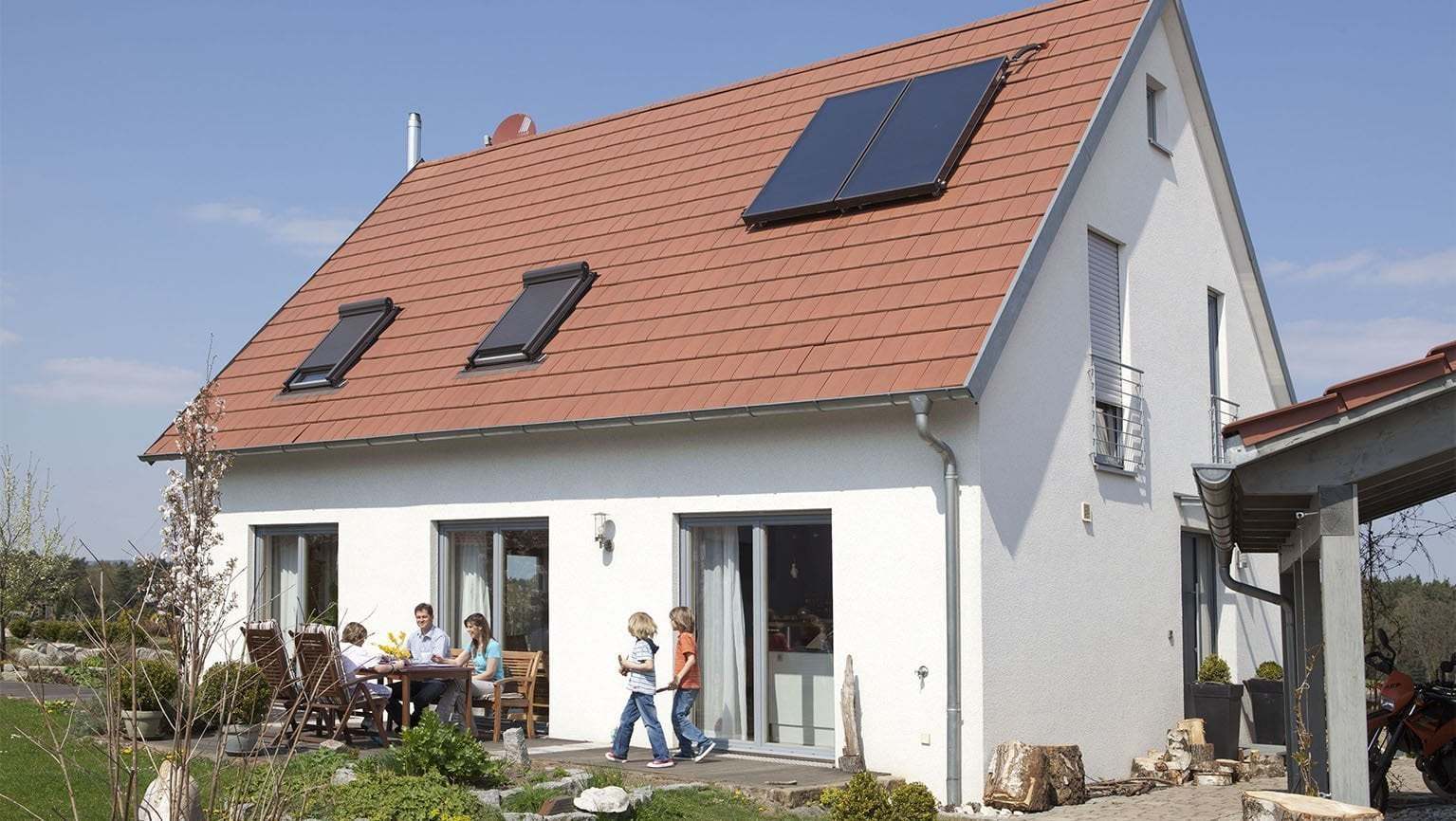 Vor einem Einfamilienhaus mit einer Photovoltaik-Anlage sitzt eine Familie an einem Tisch auf der Terrasse.