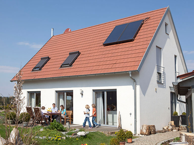 Vor einem Haus mit einem Solarmodul auf dem Dach sitzt eine Familie auf der Terrasse an einem Tisch.  